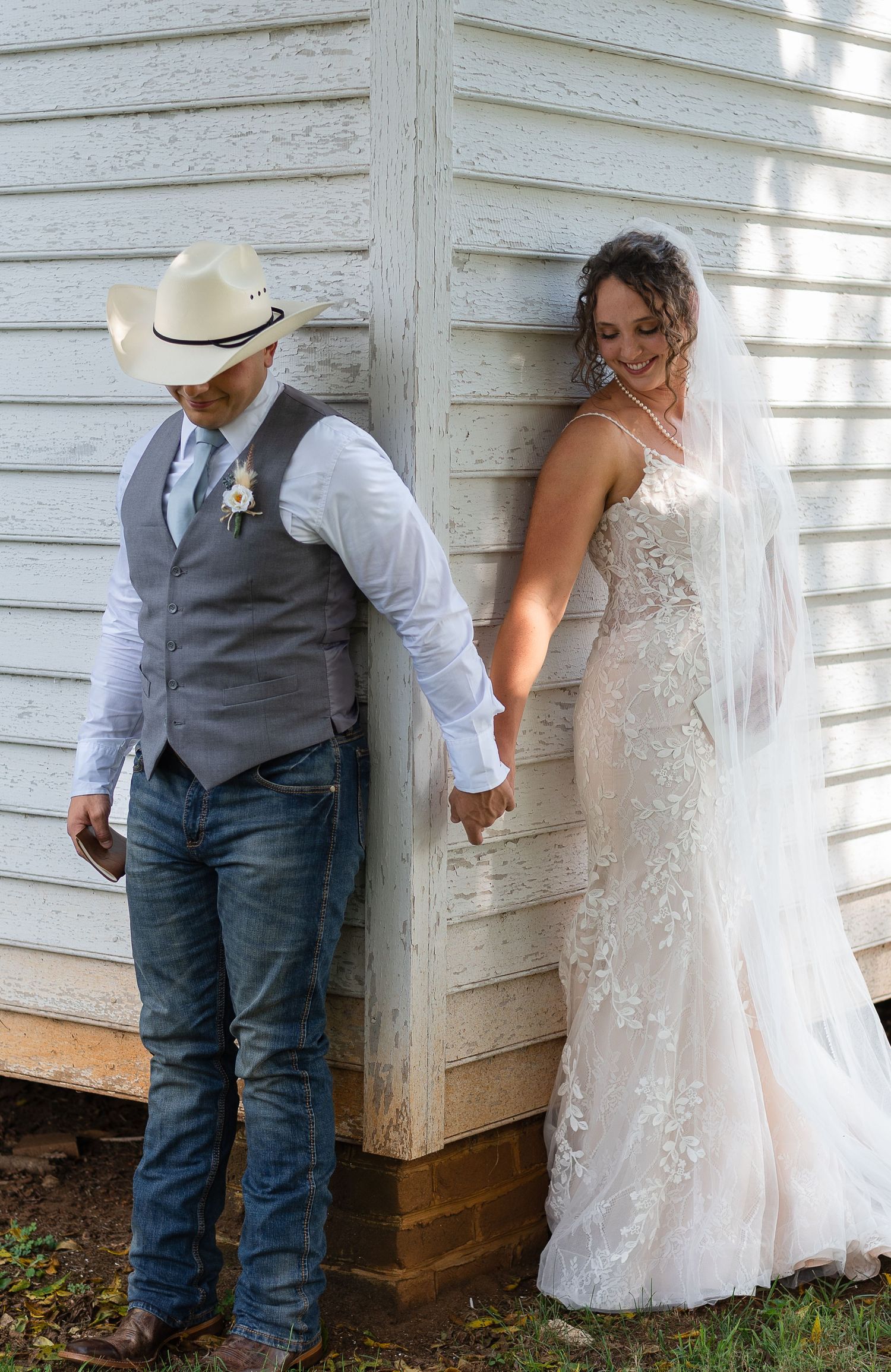 A romantic first look moment between a couple in rustic wedding attire against a white wooden barn wall.