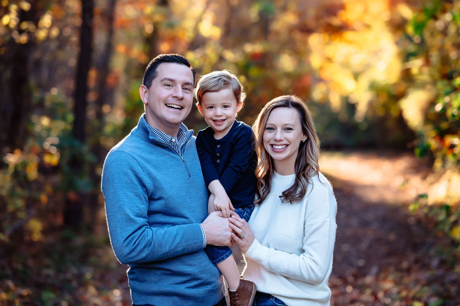 father holding son between he and mom all looking at camera smiling during dallas outdoor fall family photo session