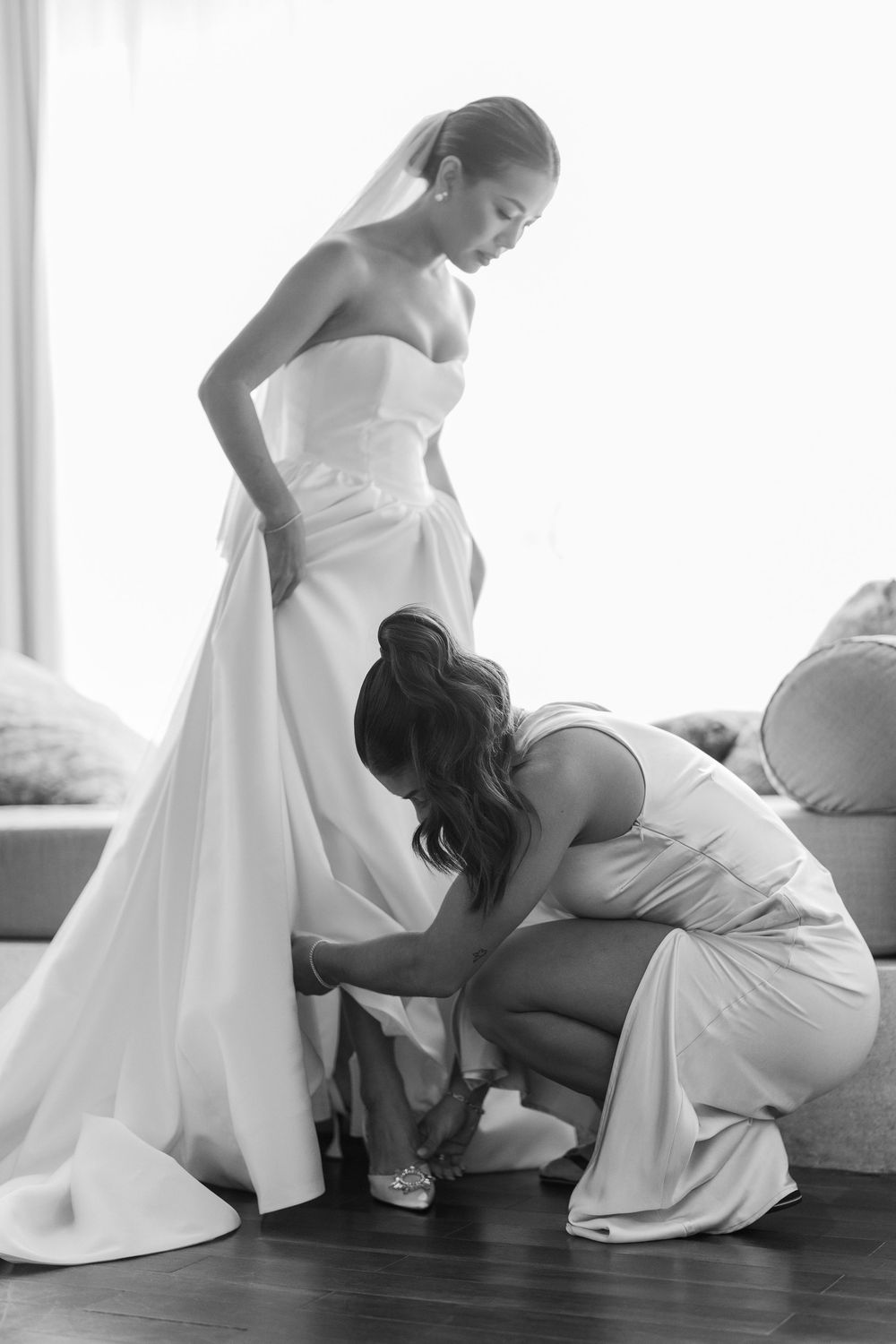 Women in white dresses help adjust wedding gown details in a bright room.