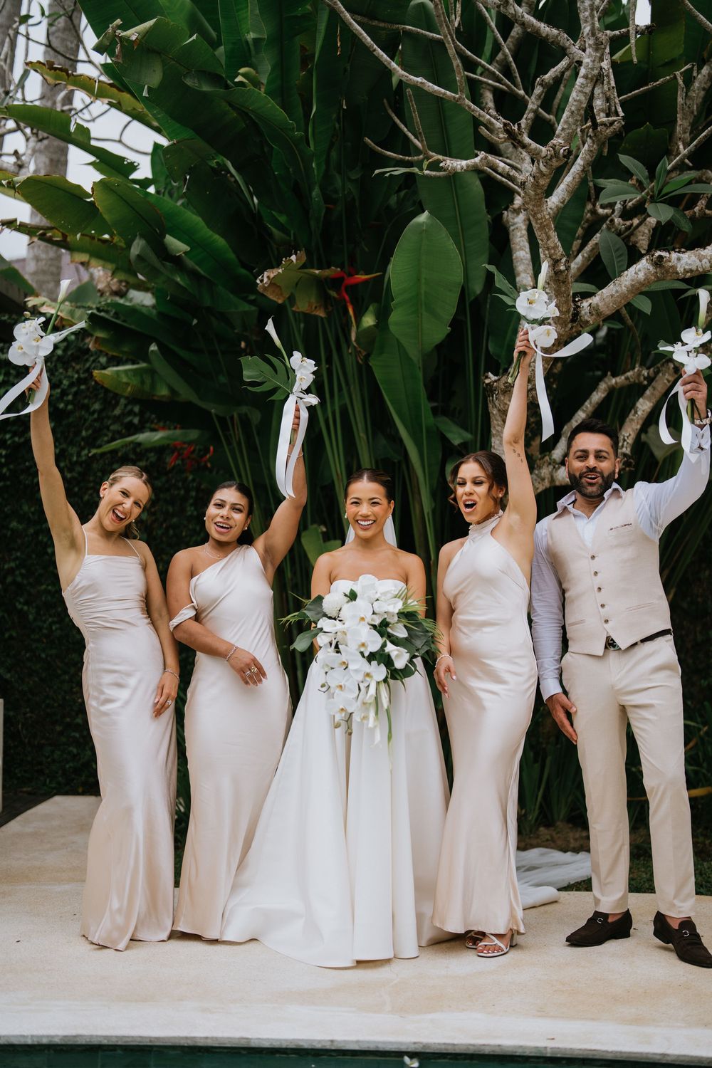 Wedding party in champagne and white attire celebrates joyfully against tropical foliage.