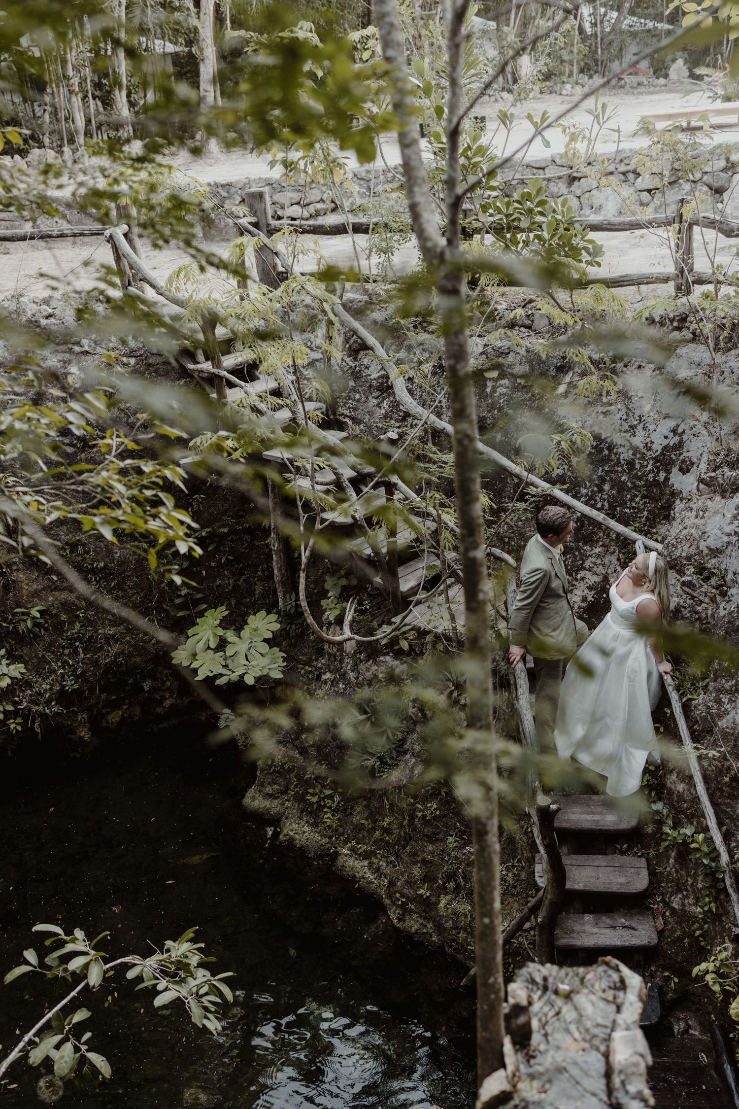 White wedding dress glimpsed through trees beside natural water feature.