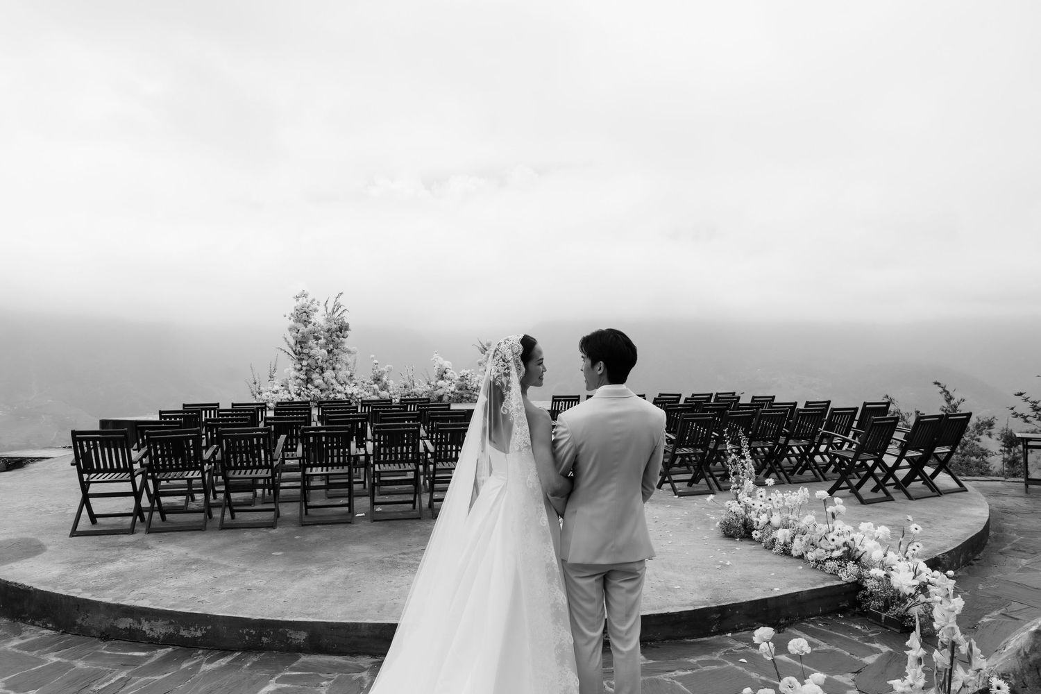 Black and white photo of bride and groom viewing empty wedding ceremony chairs set on mountain overlook.