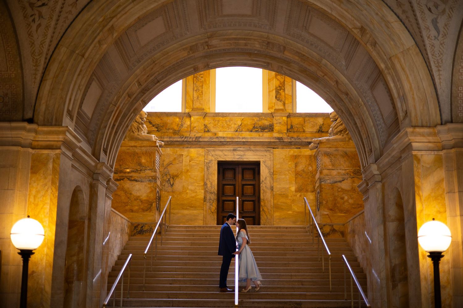 An elegant marble staircase and arched stone hallways in a historic building illuminated by warm glowing lamps.