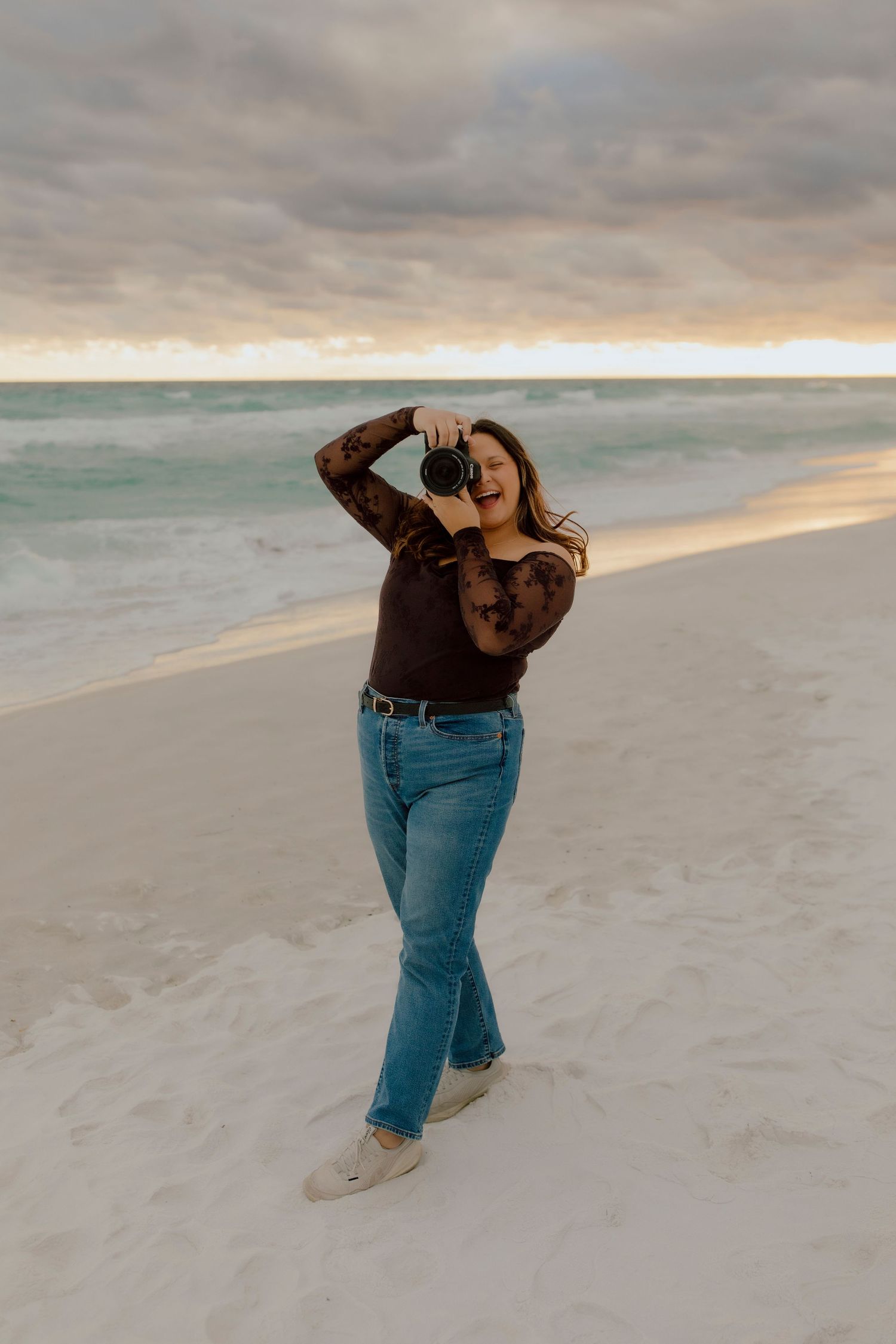 Woman in lace top and jeans photographing with camera on white sand beach at cloudy sunset.