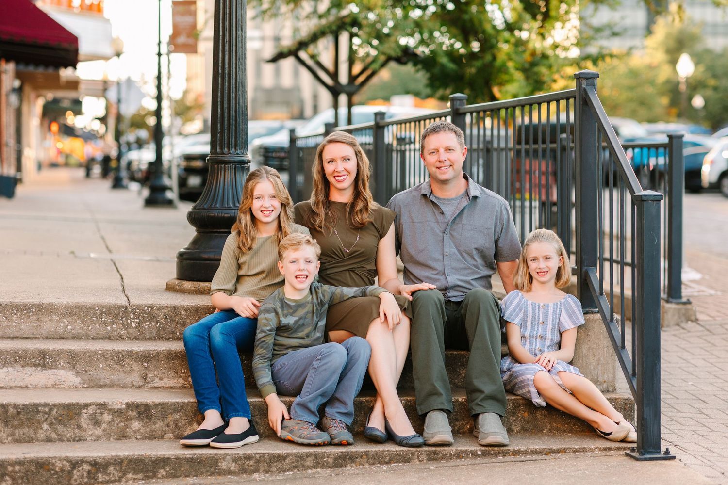 A family of five sits together on outdoor stone steps along an urban sidewalk during golden hour.