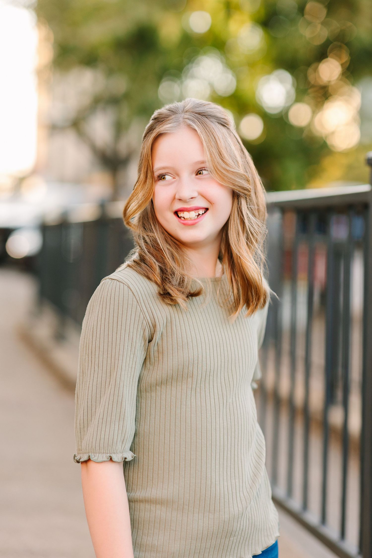 A person in a beige ribbed sweater poses outdoors with a joyful smile against a blurred background.