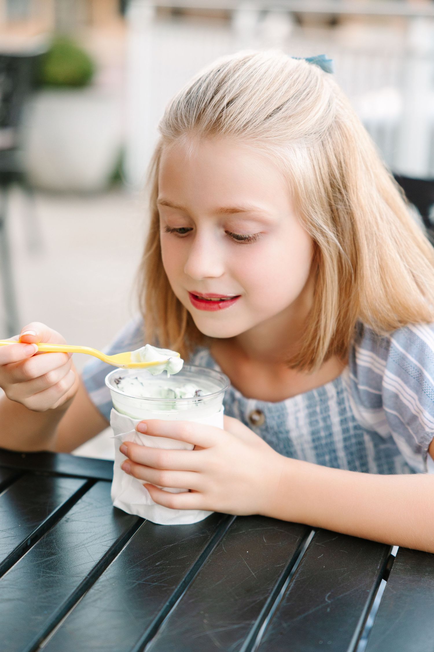 A young person wearing a blue dress smiles while enjoying yogurt or ice cream at a dark wooden table.