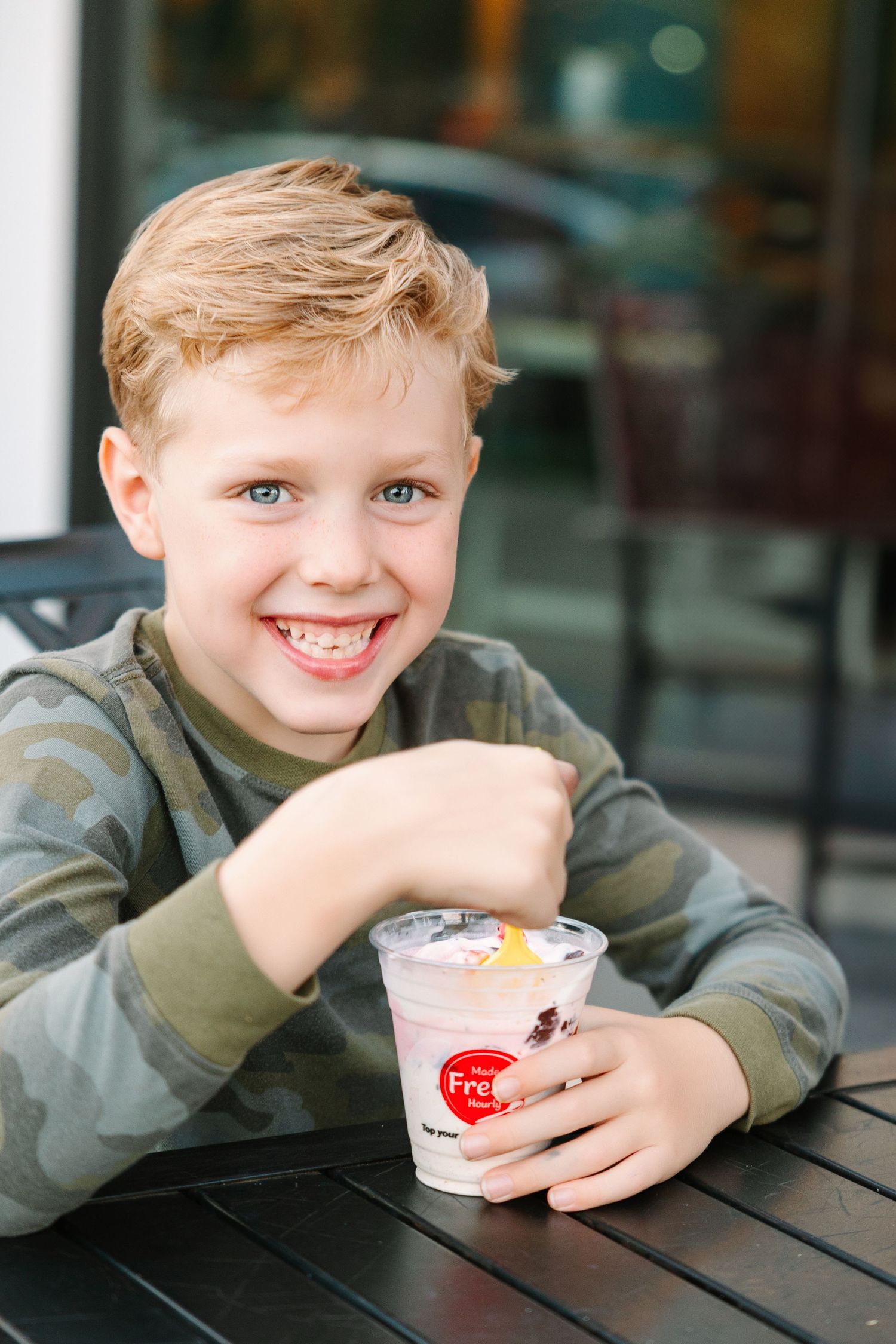 A child sits at an outdoor table enjoying a cold dessert treat with whipped cream and a cherry on top.