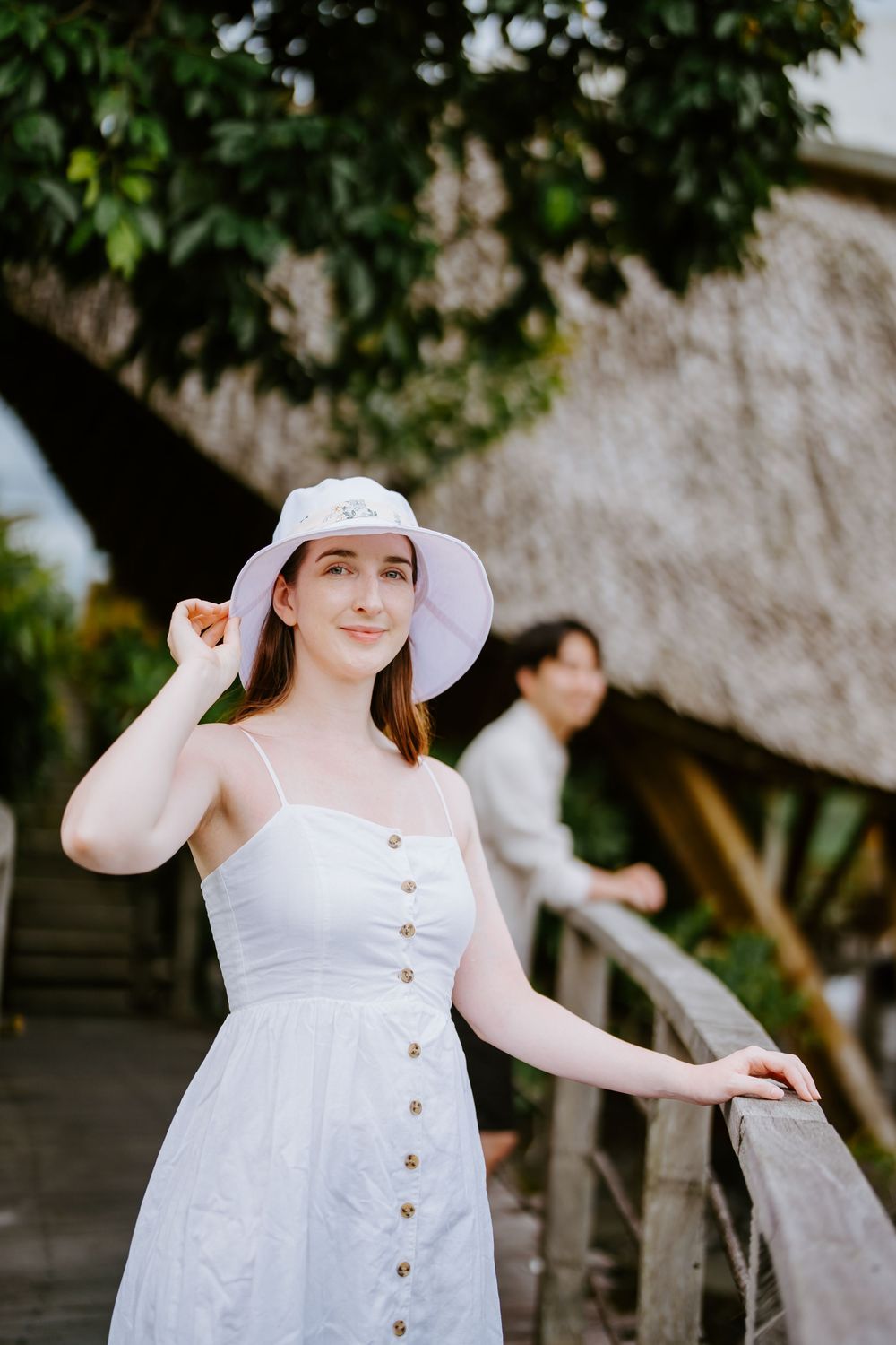 A person in a white sundress and hat poses playfully on a wooden deck with tropical scenery.