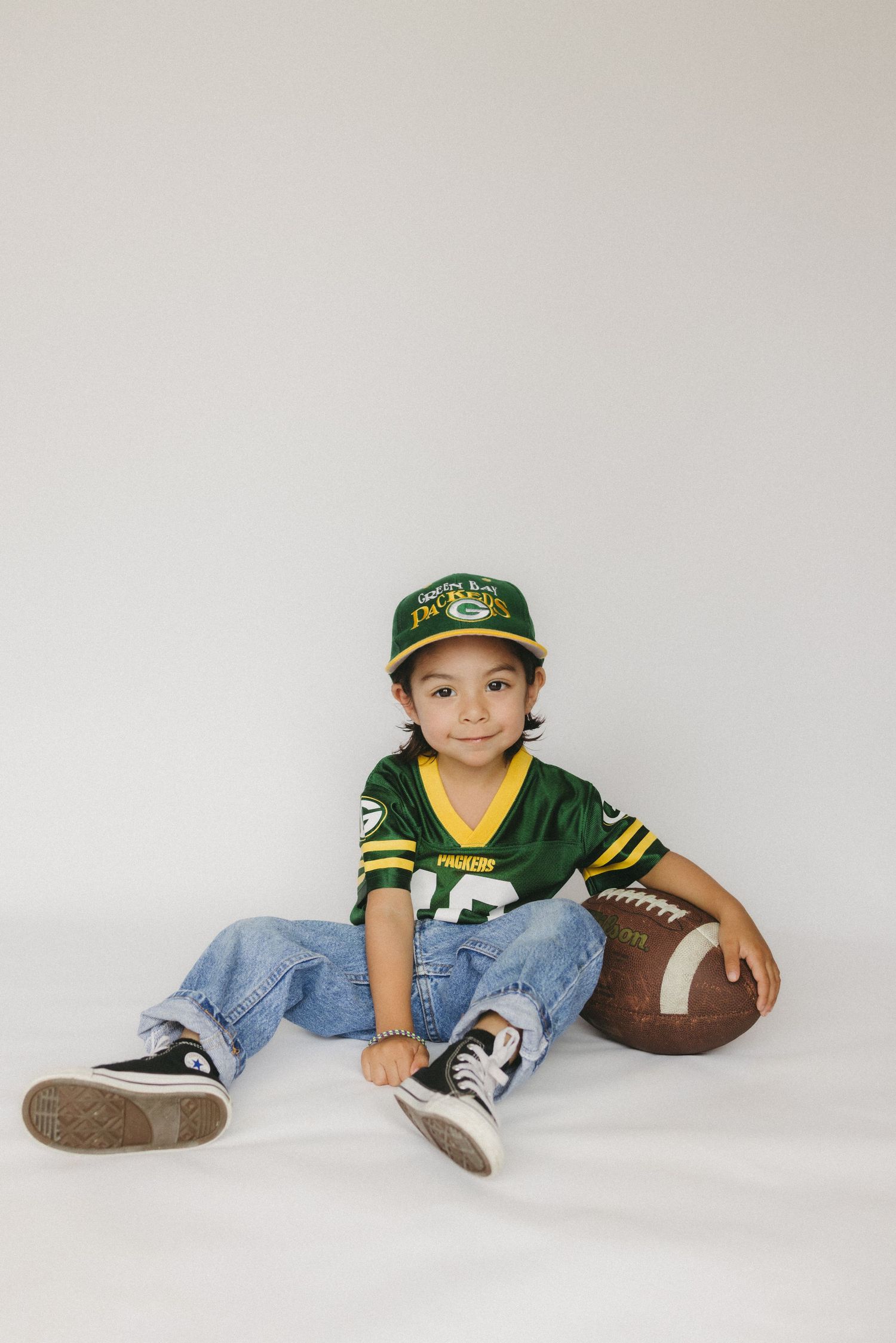 Athletic child poses on white studio backdrop with baseball cap and football.