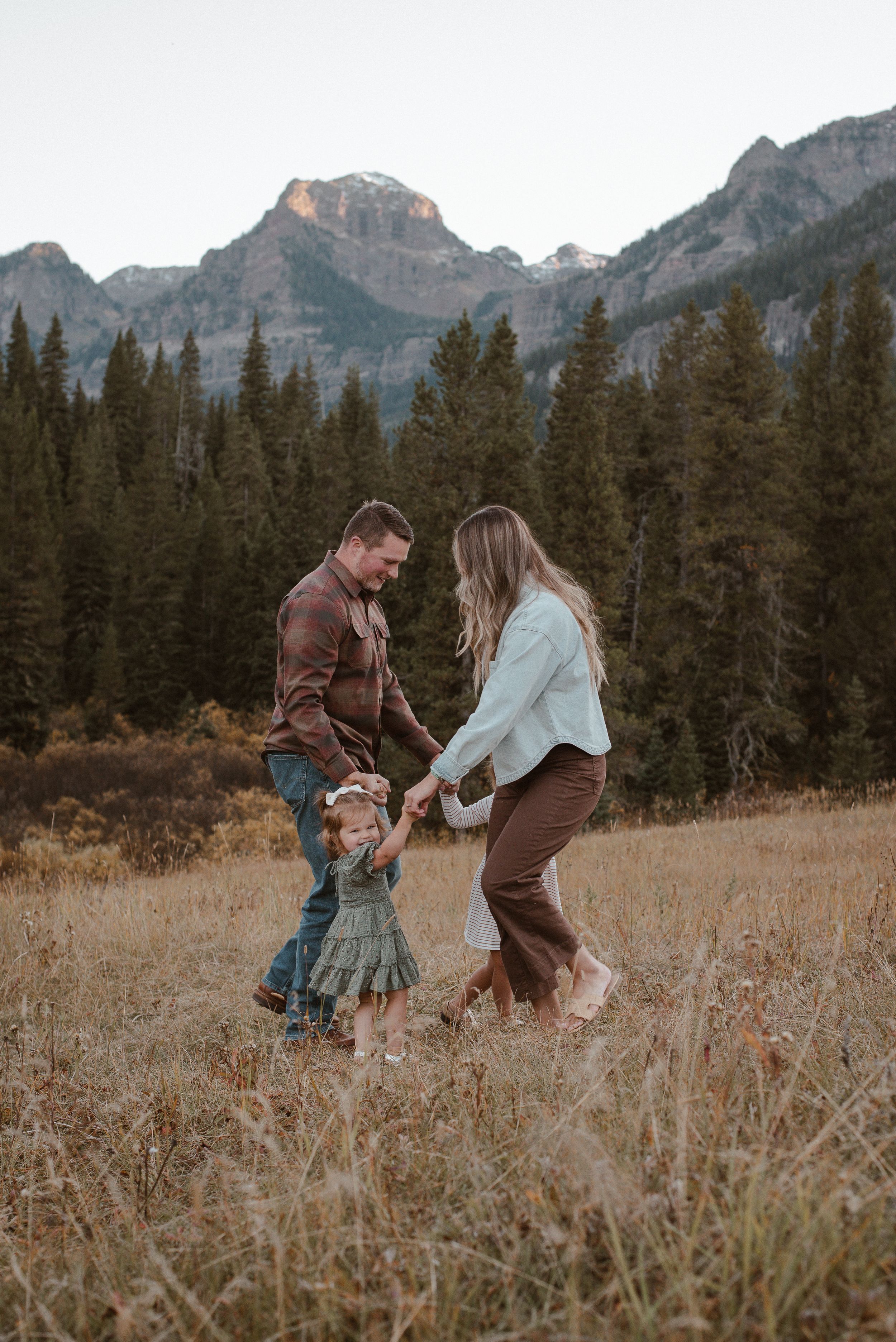 A playful family moment captured in a mountain meadow during sunset with evergreen trees in background.