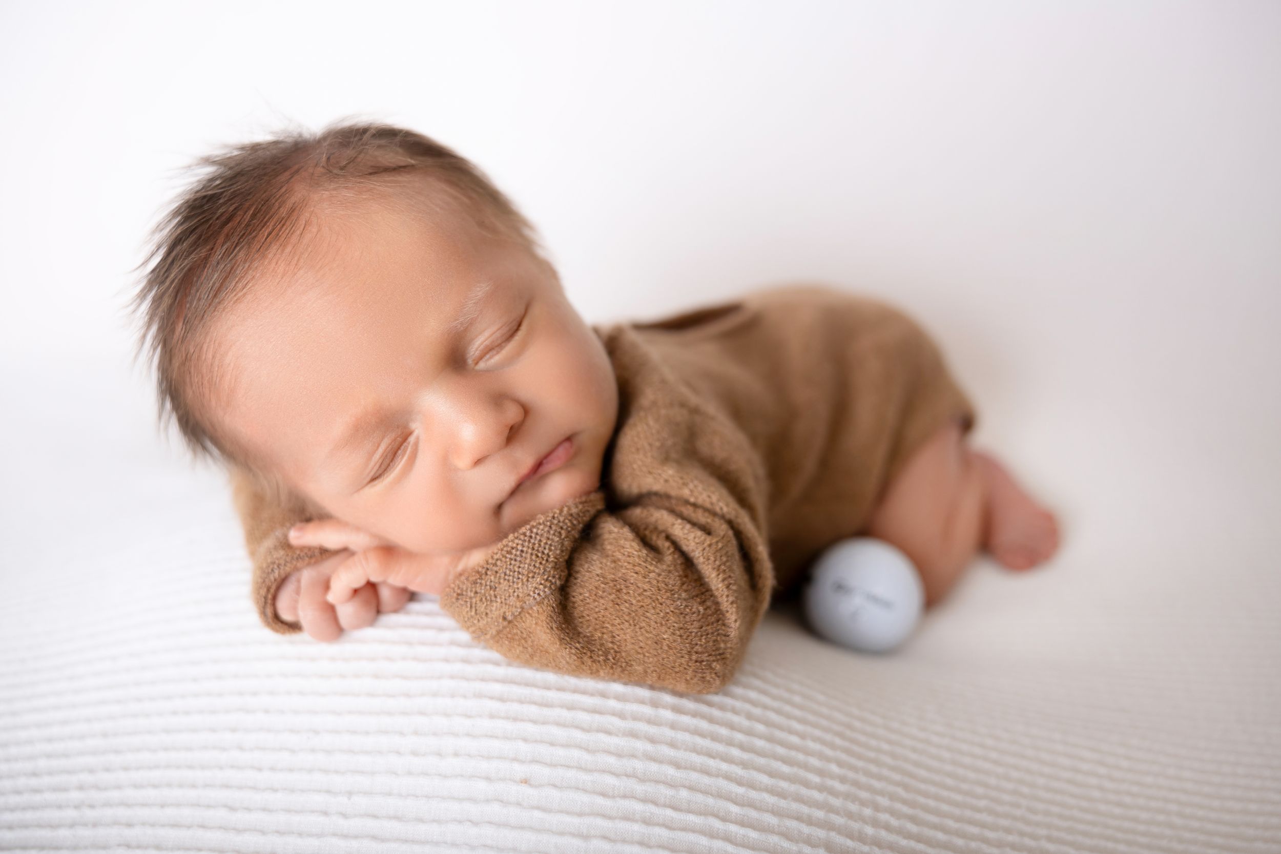 Peaceful newborn baby sleeping on white textured blanket wearing cozy brown sweater.