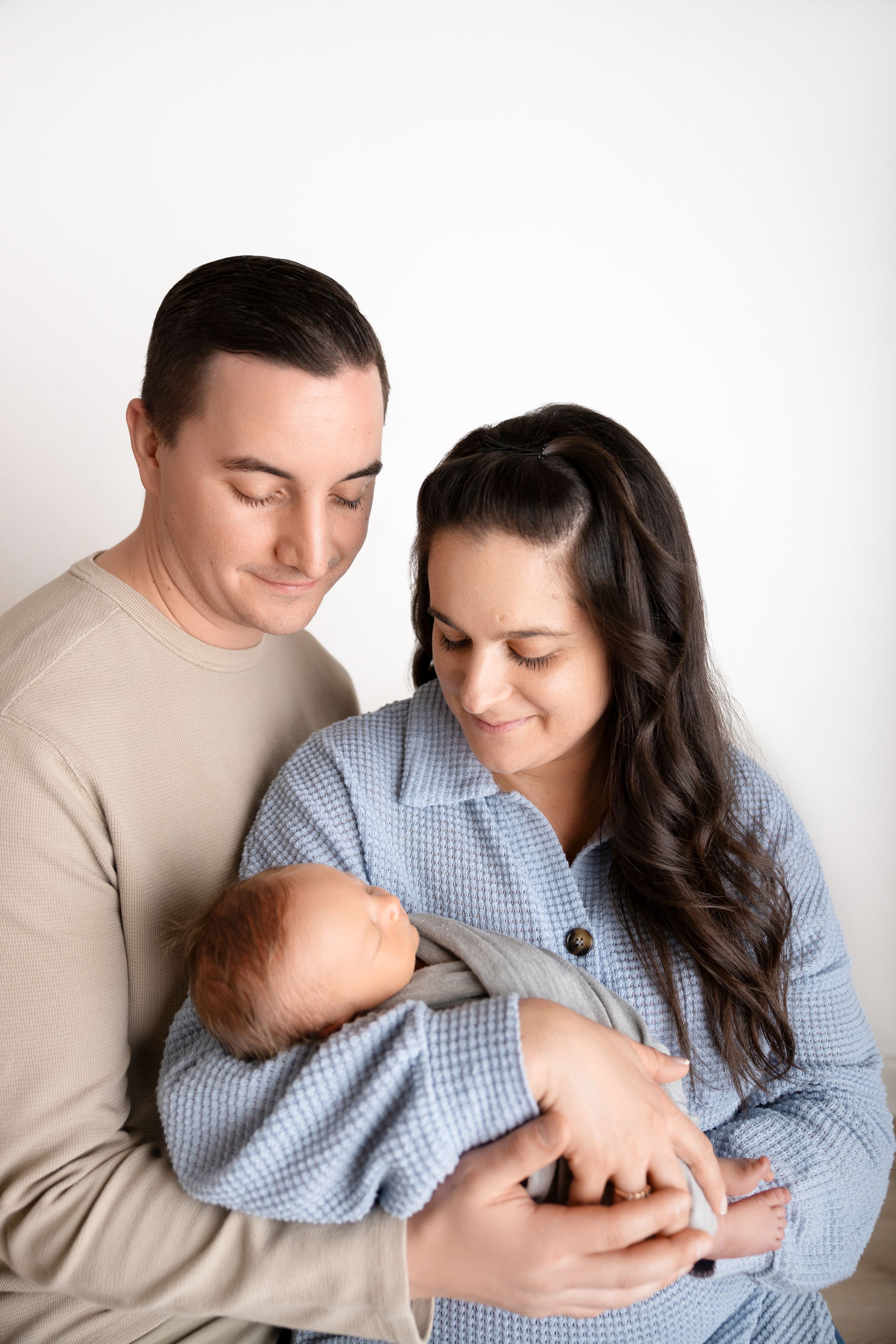 A couple in casual attire gazes lovingly at their newborn baby against a white background.