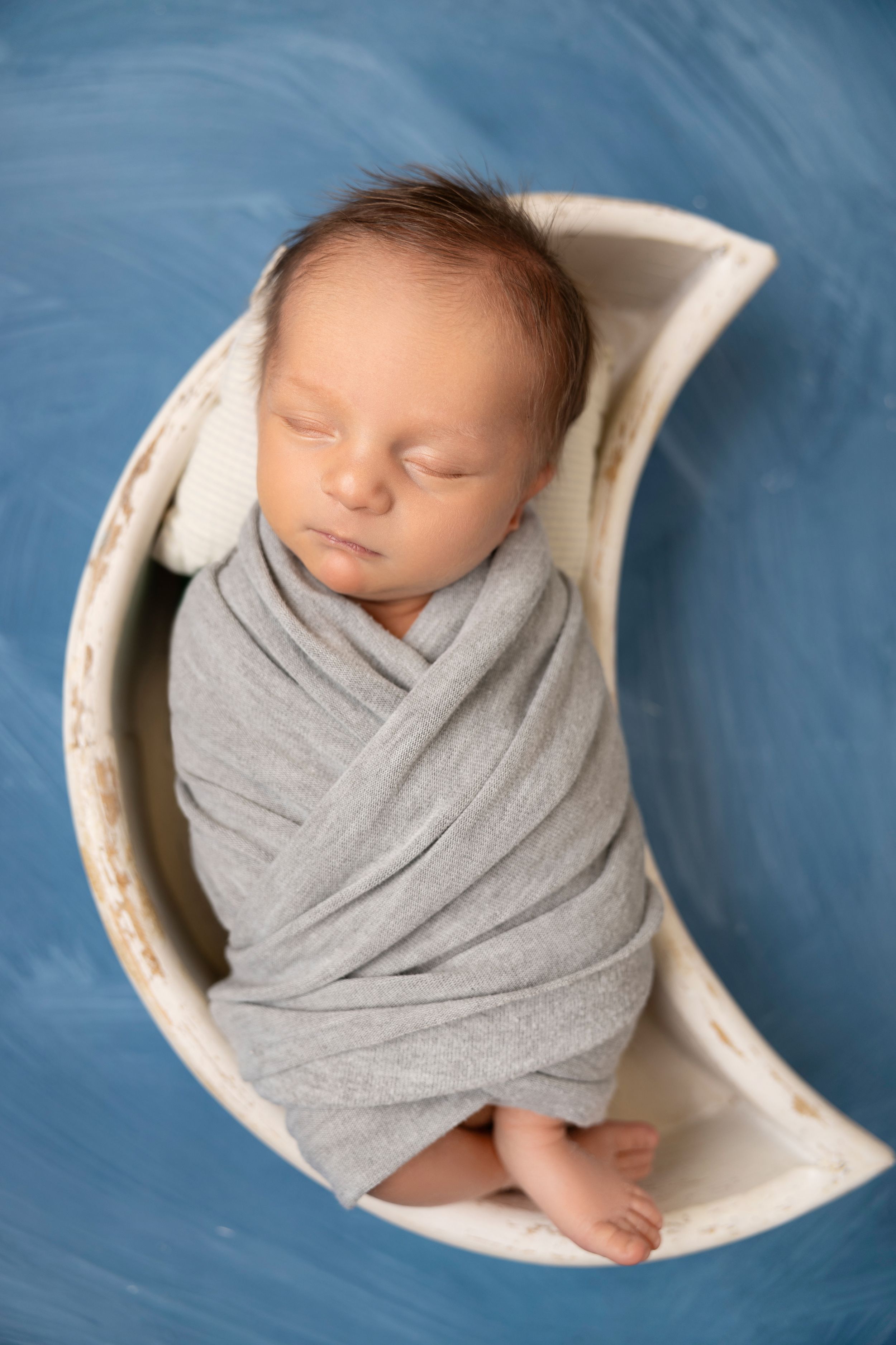 Sleeping newborn baby wrapped in soft gray swaddle blanket nestled in white round bowl against blue backdrop.