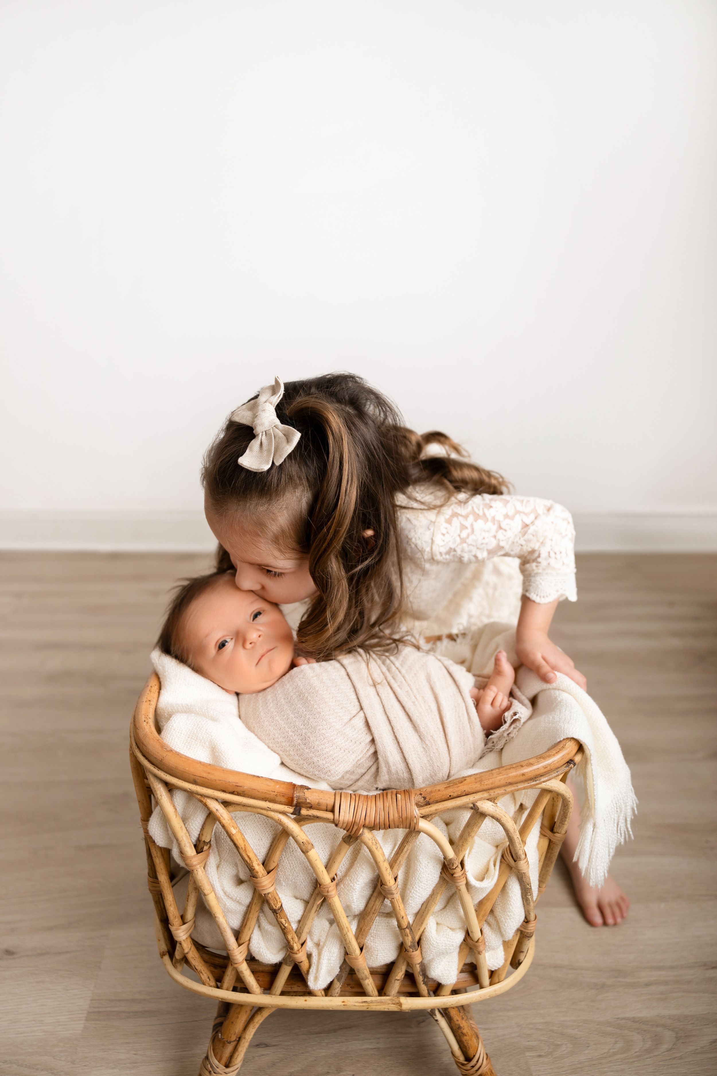 A baby lies peacefully in a vintage rattan basket on a wooden floor while wearing a cream-colored outfit.