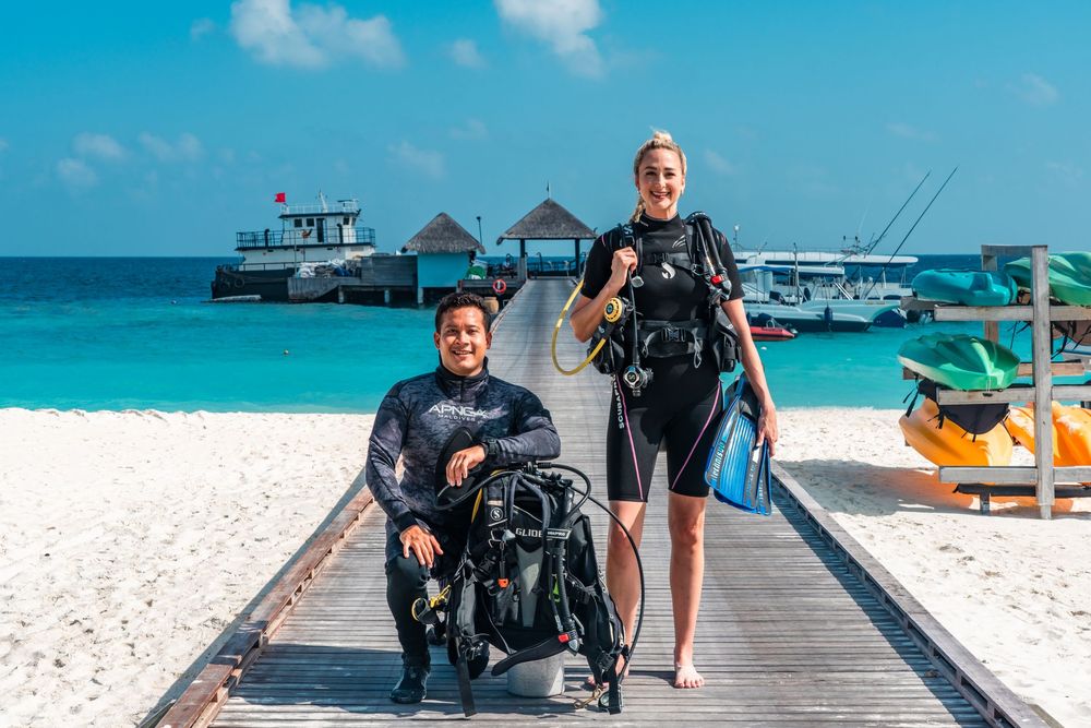 Beachgoers and scuba diving equipment on a wooden walkway with crystal clear turquoise waters in the background.