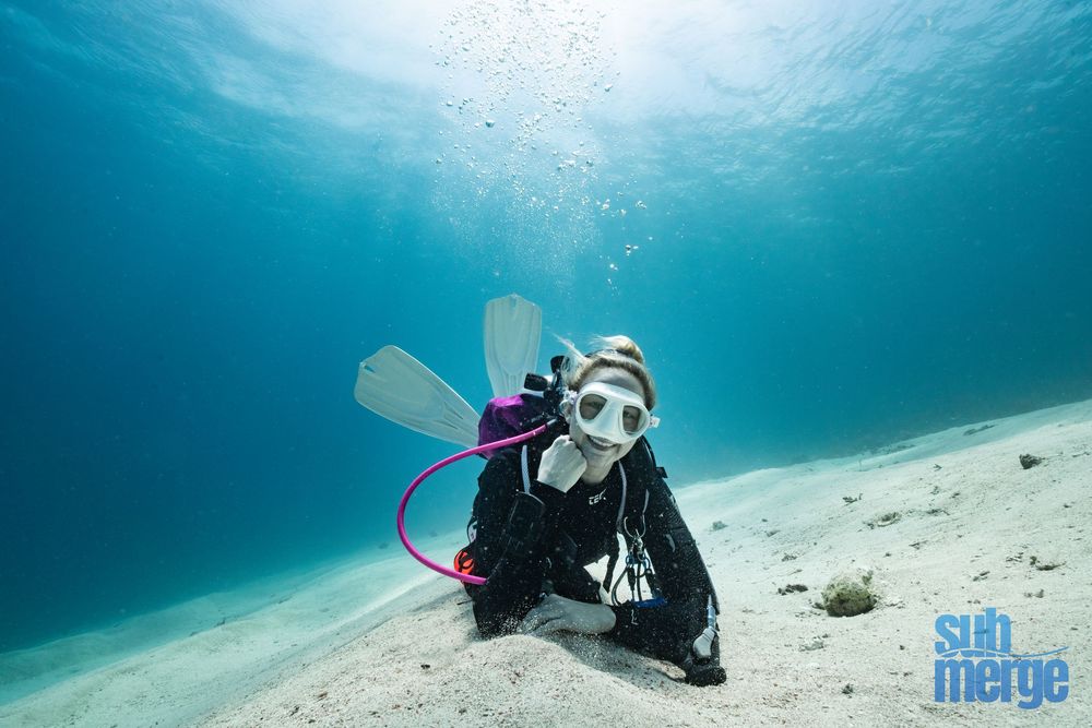 A scuba diver swims along the sandy ocean floor in crystal clear turquoise waters.
