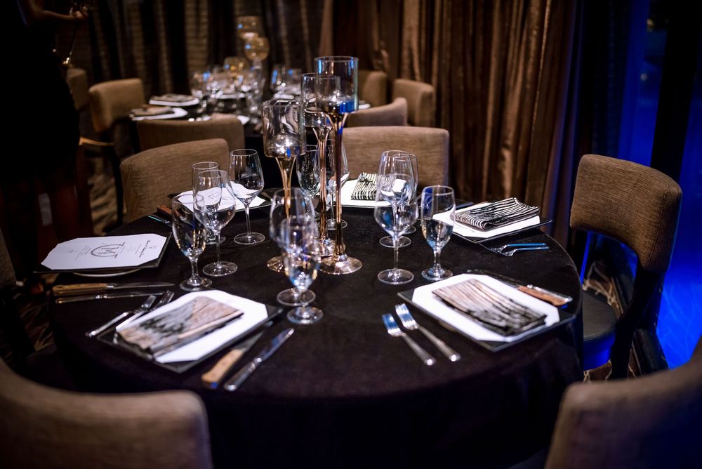 Elegant restaurant dining tables set with wine glasses and white linens in a dimly lit room with blue accent lighting.
