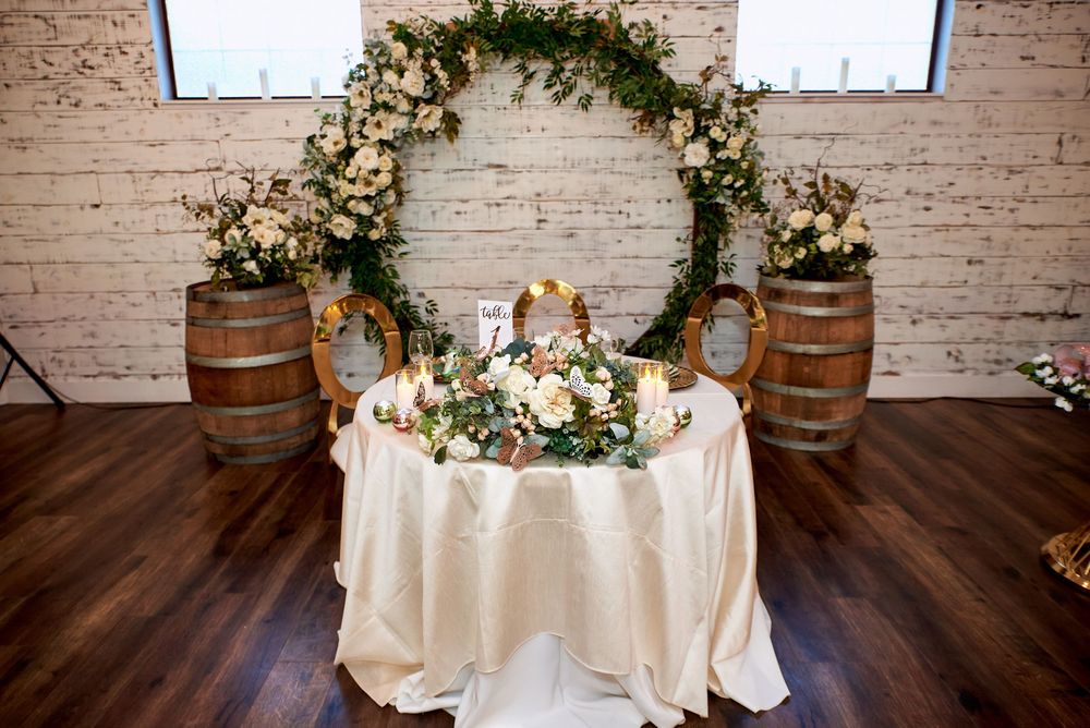 Wedding reception sweetheart table with floral arch, wine barrels and white tablecloth in rustic indoor venue.