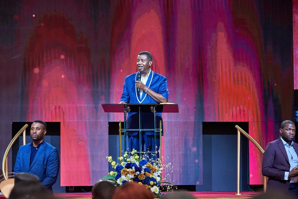 Speaker at a podium delivers remarks on stage with dramatic red and purple lighting and floral decorations. Pastor E.A Adeboye in Houston TX