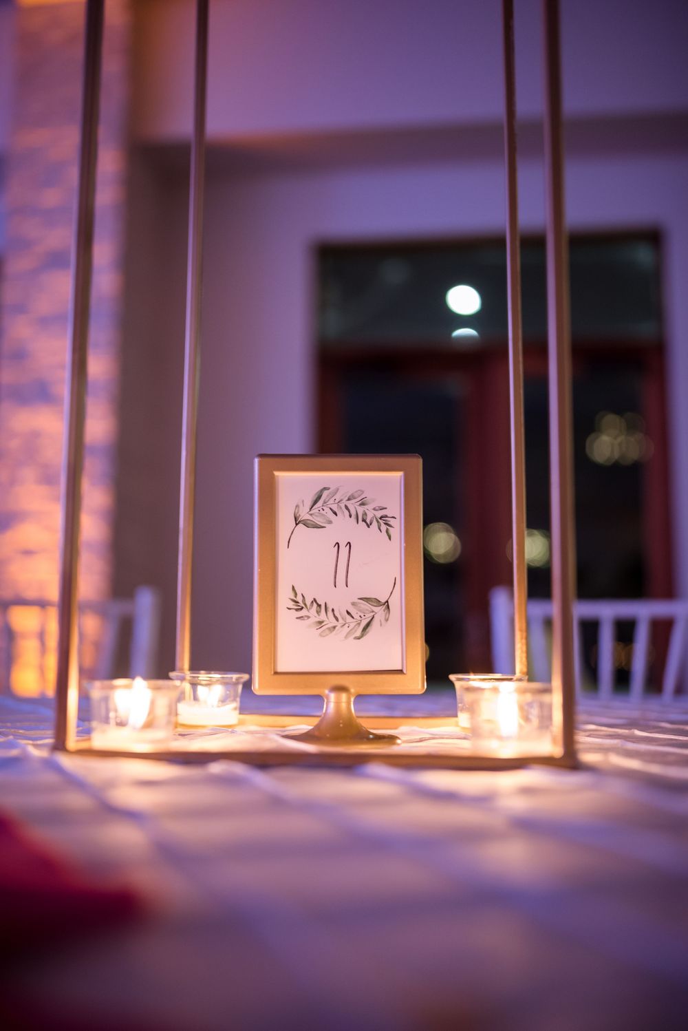 A framed table number card sits elegantly between glowing candles at a wedding reception with purple ambient lighting.