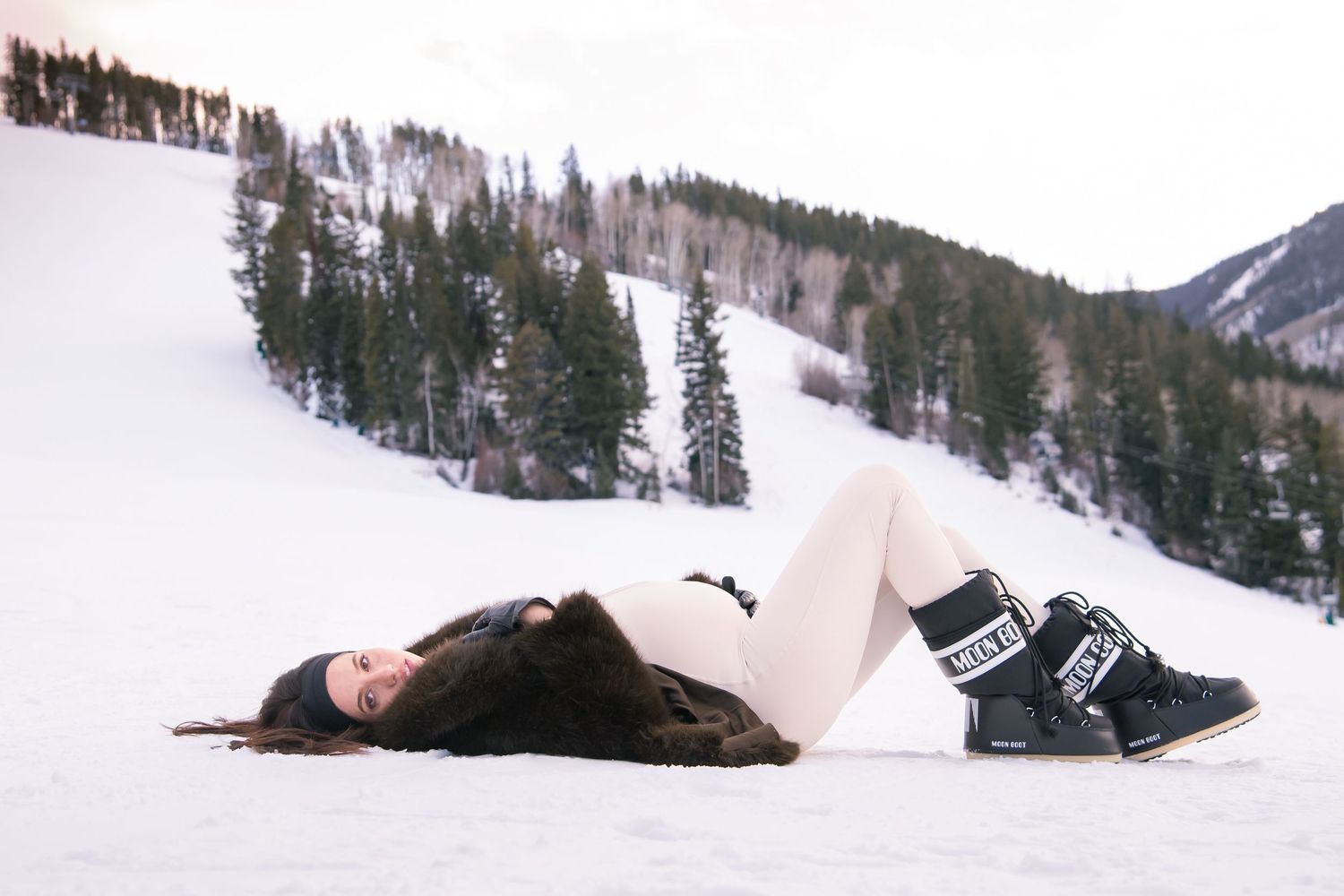 A cute stylish woman lays in the snow with mountains in the background for her winter fashion maternity photos in Vail Colorado