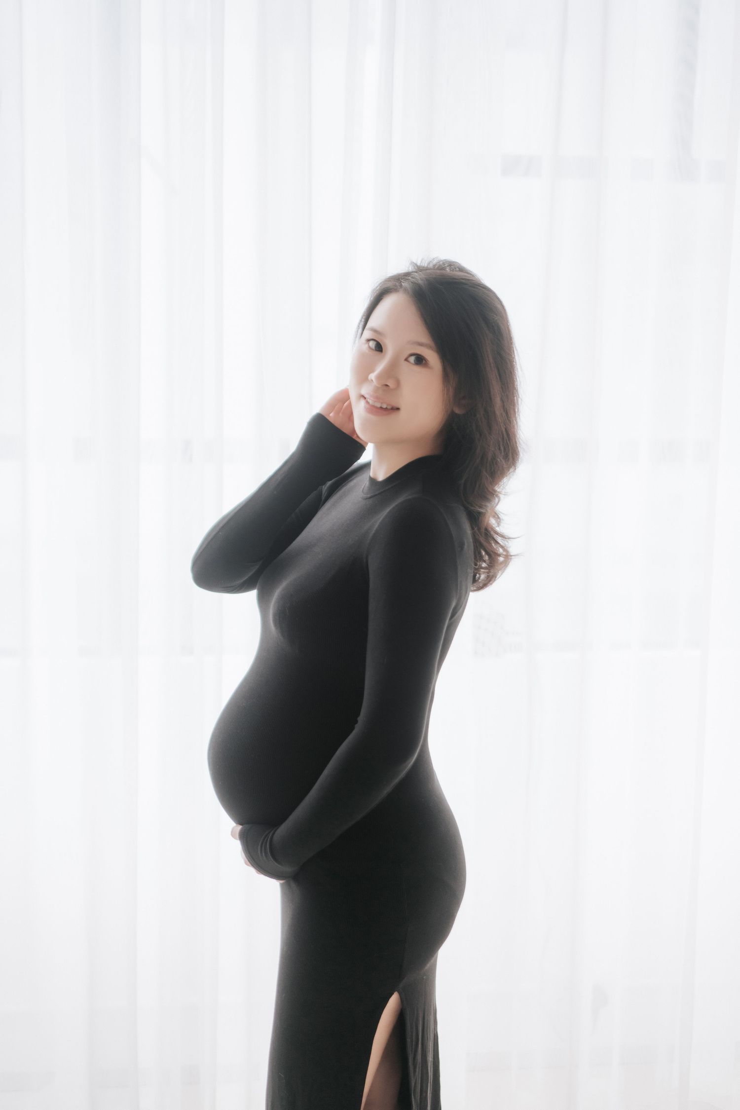 A pregnant woman in a long black dress poses gracefully against a white curtain backdrop in soft lighting.