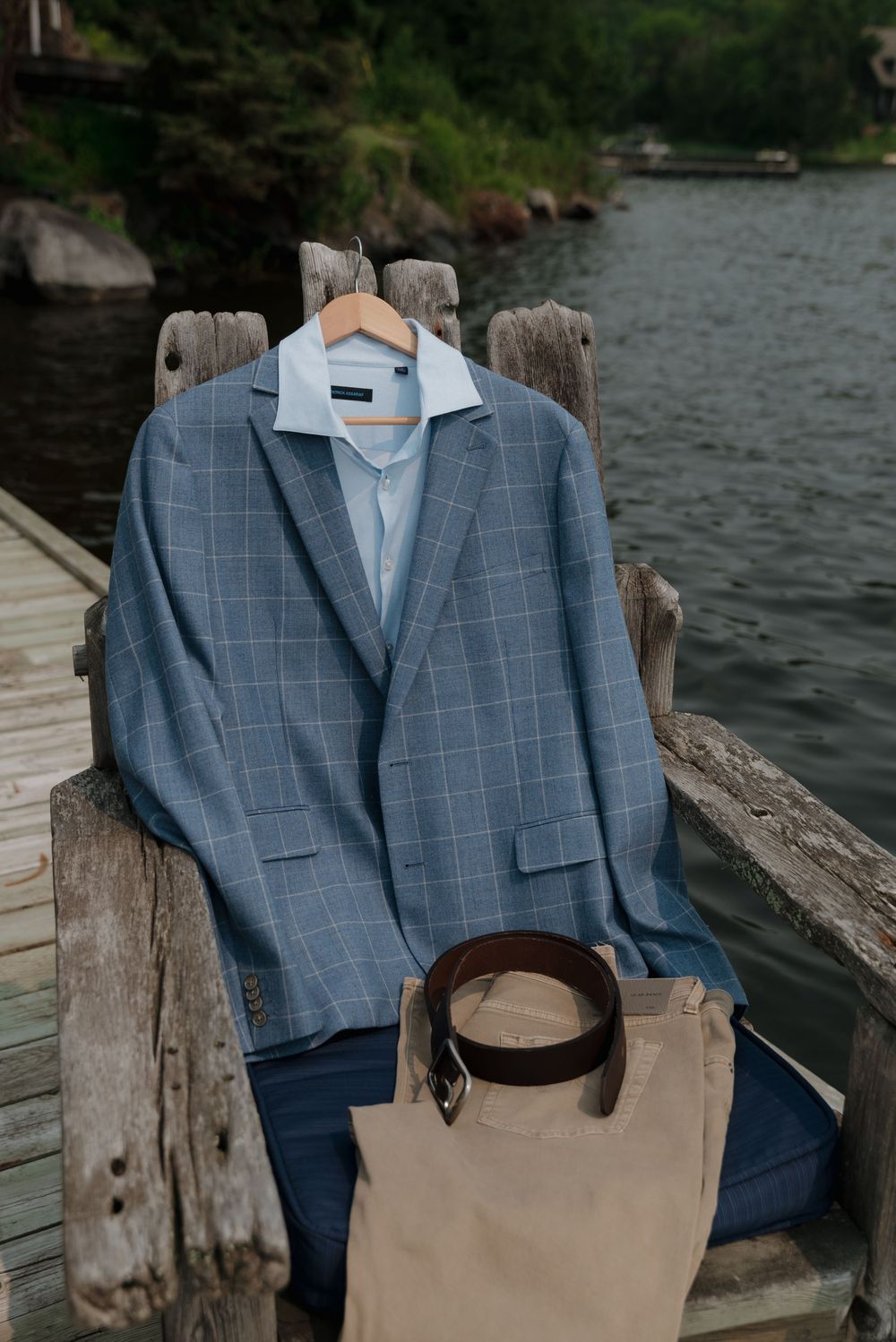 Blue checkered suit jacket and tan bag displayed on wooden dock railing with lake view.