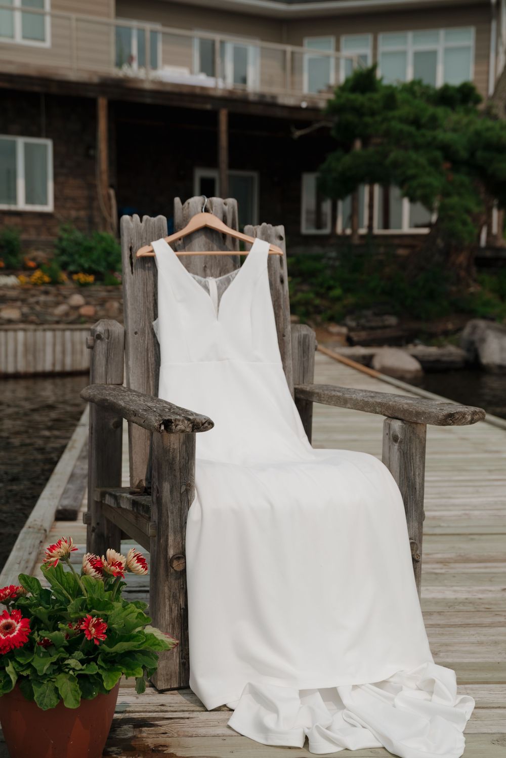 White wedding dress displayed on wooden dock with red flowers and lake in background.