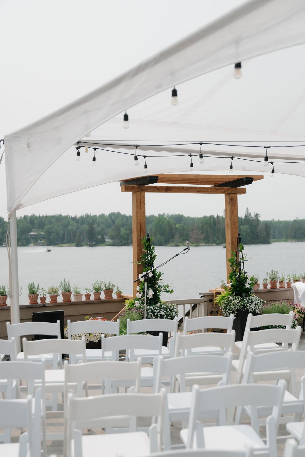 White folding chairs arranged under a tent canopy overlooking a serene lakefront setting.