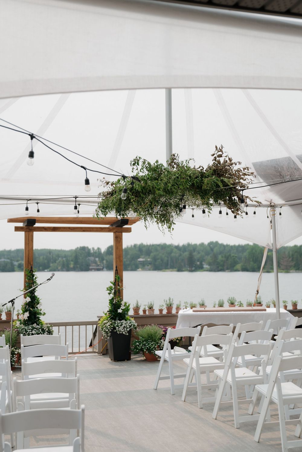 White chairs arranged under a tent canopy with greenery-decorated wooden arch overlooking a waterfront.