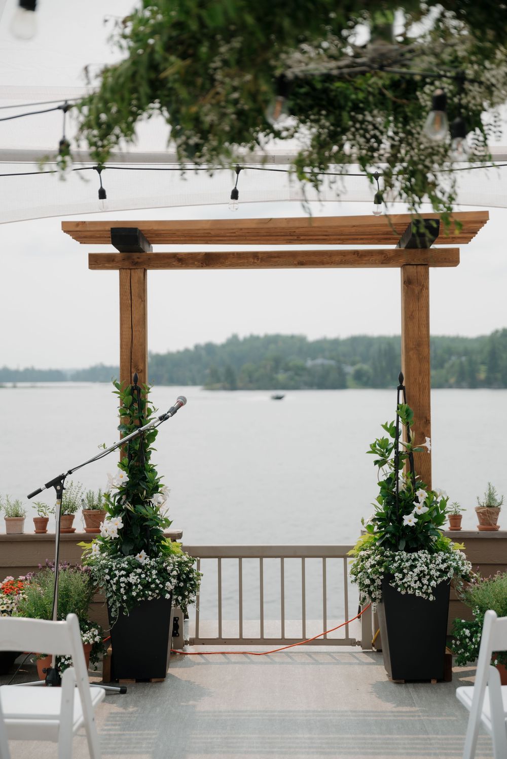 Wooden pergola decorated with white flowers and greenery overlooks a serene lake view from a deck.
