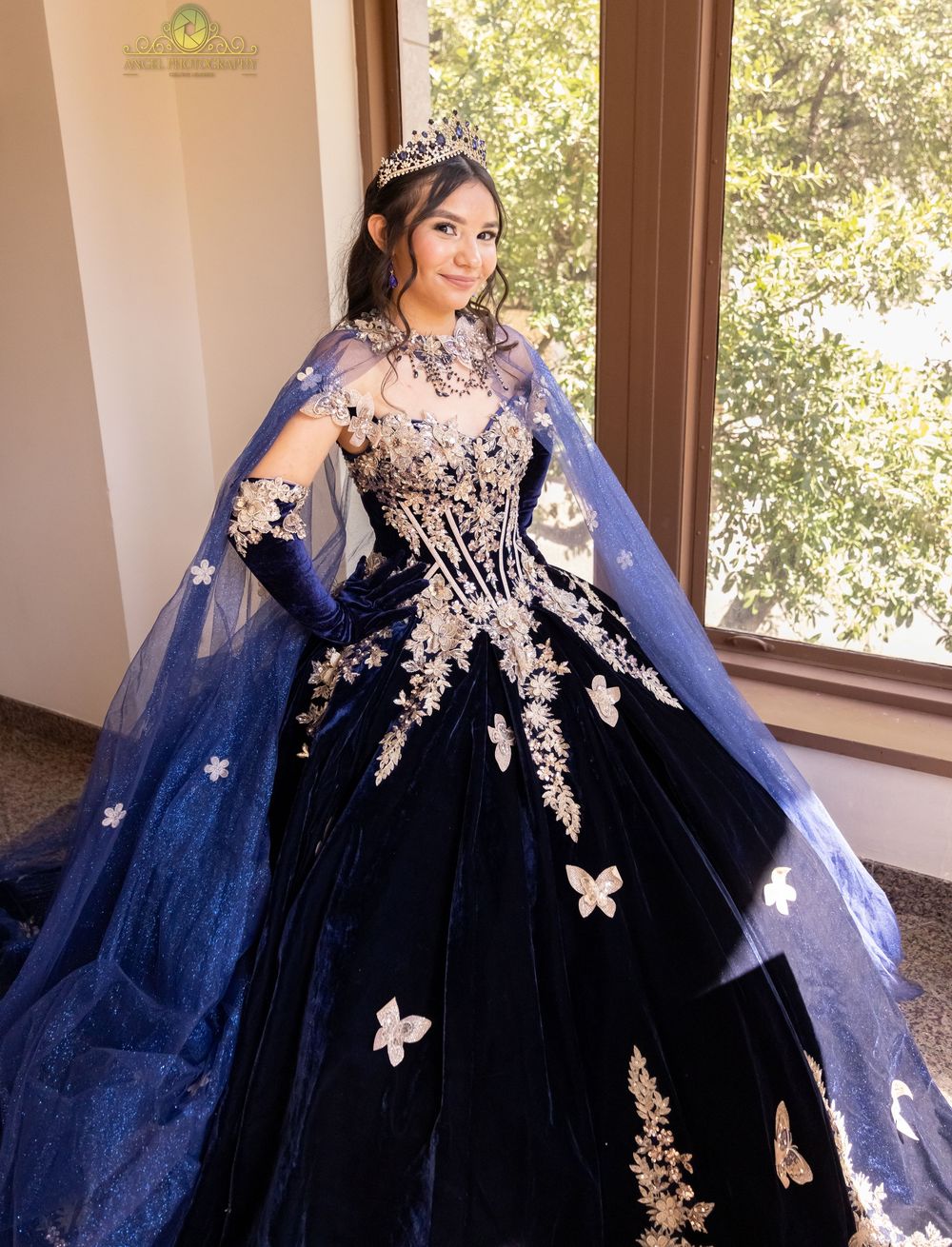 Elegant navy blue ball gown with intricate gold lace appliques and flowing cape showcased in a sunlit room.