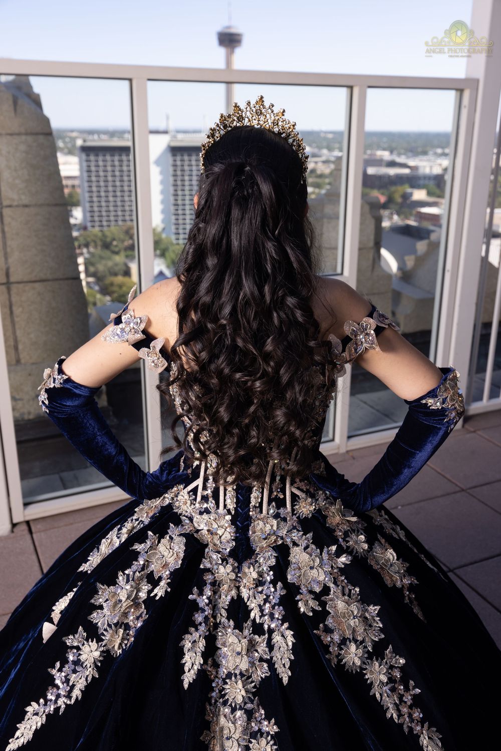 Person wearing a navy blue gown with intricate silver embellishments stands by large windows overlooking a cityscape.