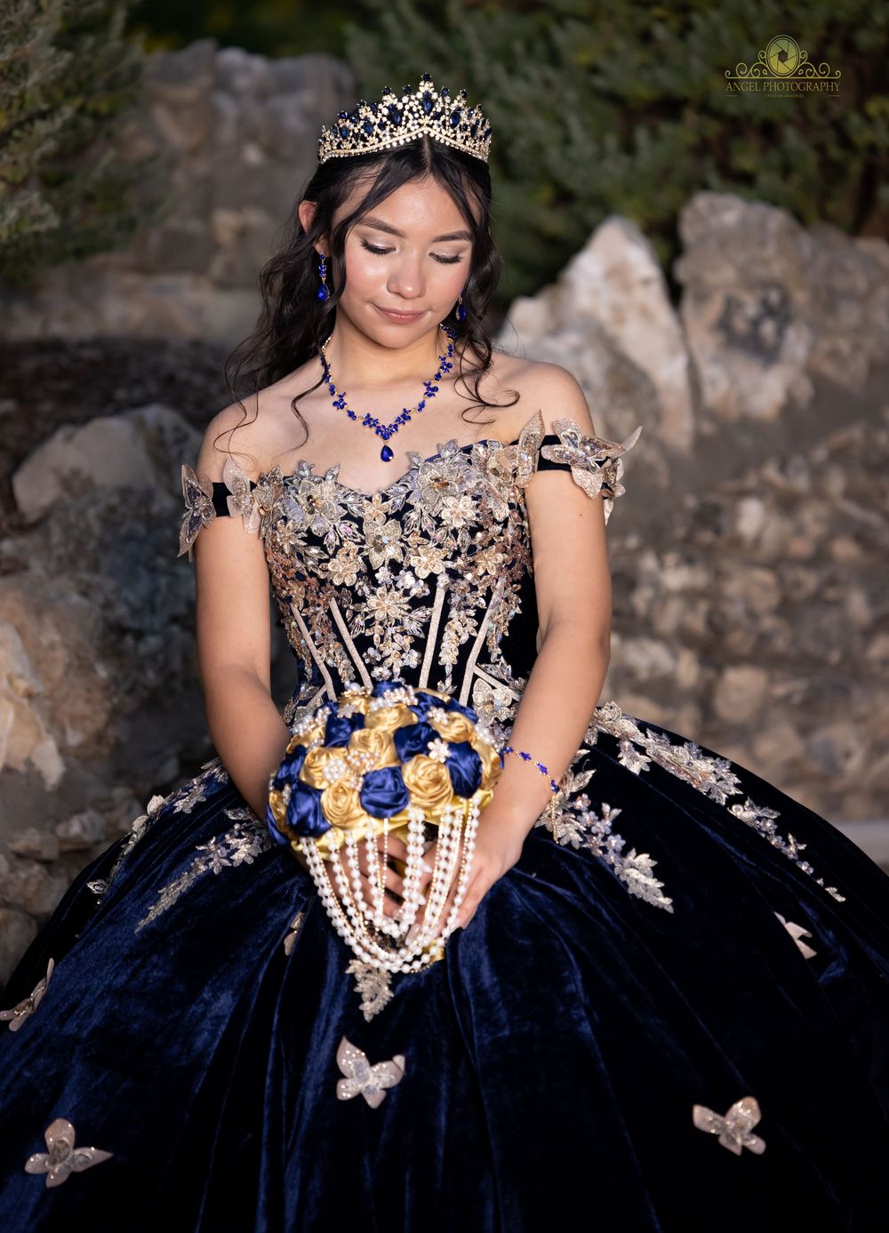 A quinceañera dress in navy blue velvet with silver embellishments, tiara and matching jewelry against a stone backdrop.