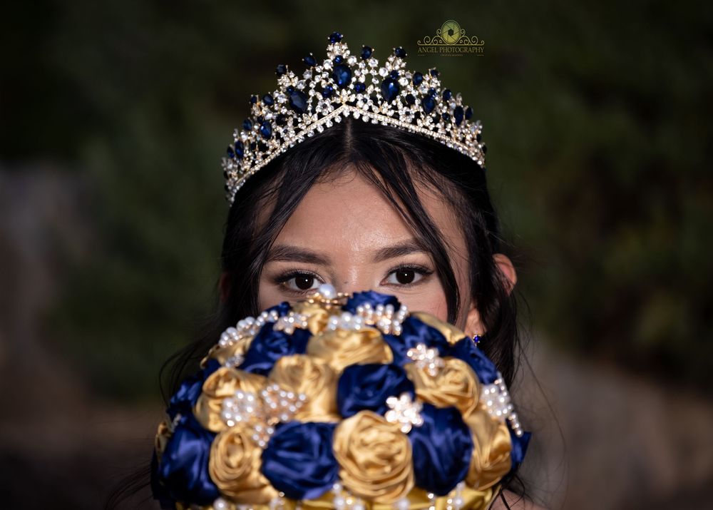 A sparkling tiara adorns dark hair above a bouquet of royal blue and gold roses held at face level.