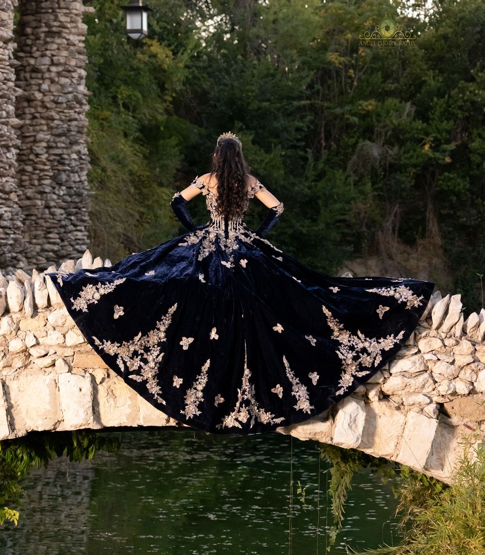 A person in a flowing black dress with butterfly wings poses dramatically against a stone wall in nature.
