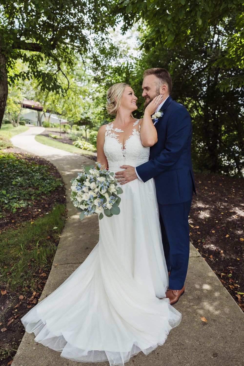 A romantic wedding couple shares a tender moment on a garden path surrounded by lush greenery and trees.