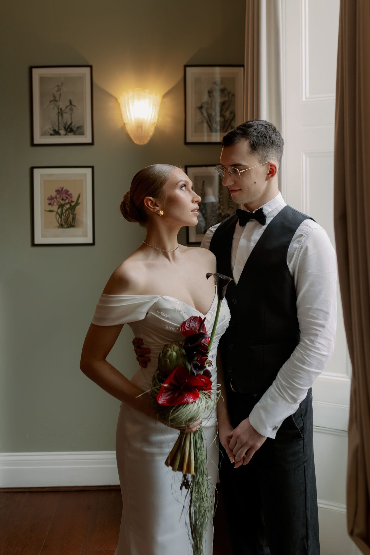 A romantic wedding couple shares an intimate moment in an elegantly decorated room with framed artwork on the walls.