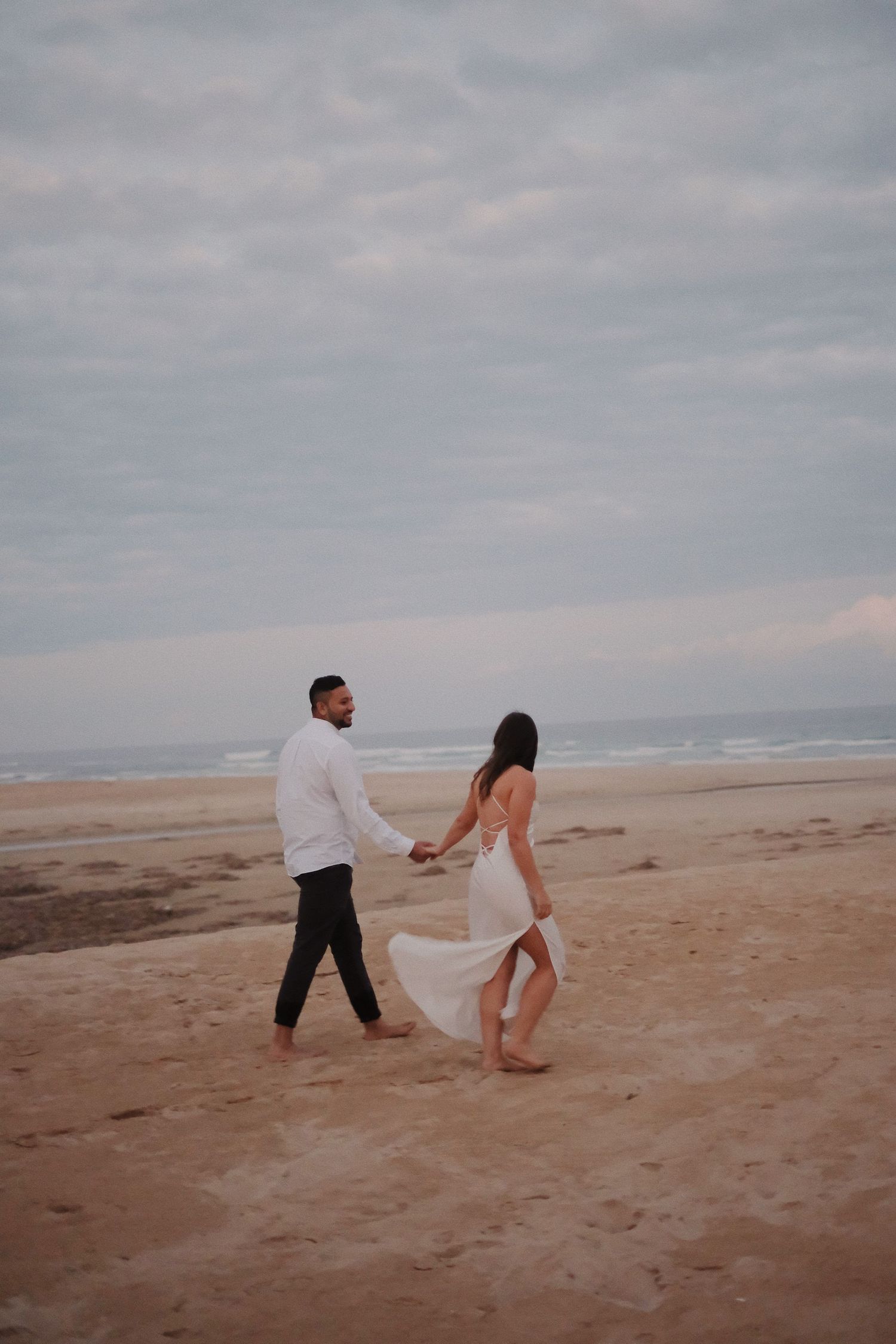 Couple walking barefoot on sandy beach holding hands, bride in flowing white backless dress under cloudy sky.