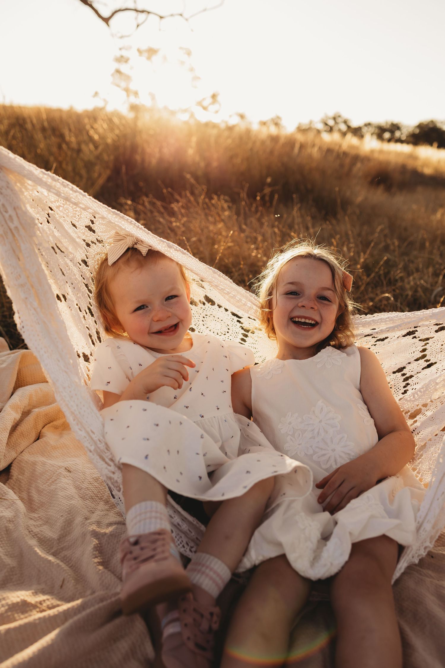Two children in white dresses laugh and play together on a blanket during golden hour sunset.