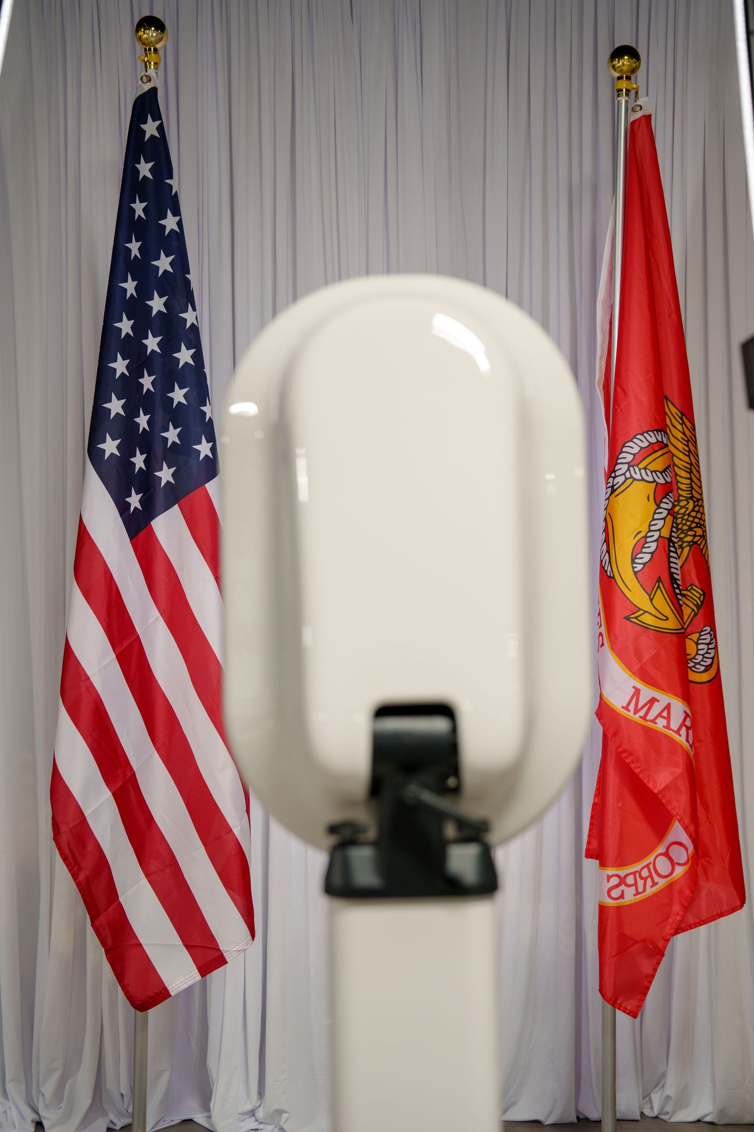 American and Marine Corps flags displayed on poles against a white curtain backdrop with round light fixtures visible.