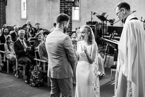 Black and white photo of bride laughing joyfully while exchanging vows with groom at church altar.