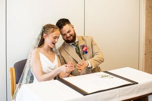 Bride and groom laughing together while signing their wedding certificate at a white table.