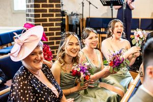 Smiling bridesmaids in sage green dresses holding colorful bouquets seated at a wedding ceremony.