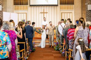 Bride and groom exchange vows at church altar surrounded by standing wedding guests in colorful attire.
