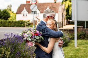 Bride embraces older man holding colorful bouquet outdoors near lavender bushes after wedding.