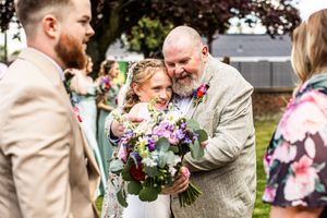 Bride hugs smiling older man with floral boutonniere while guests celebrate outdoors after ceremony.