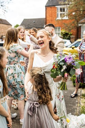 Laughing bride holds bouquet during outdoor confetti celebration with flower girl and wedding guests.
