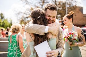 Groom in tan suit shares warm embrace with bridesmaid holding bouquet outside sunny wedding venue.